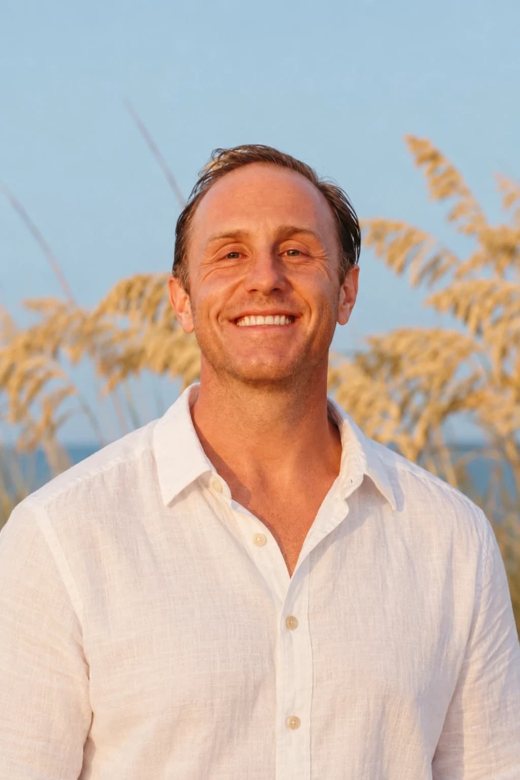 Dr. Daniel Tagge in a white linen shirt at the shore, sea oats and a pale sky behind him, smiling