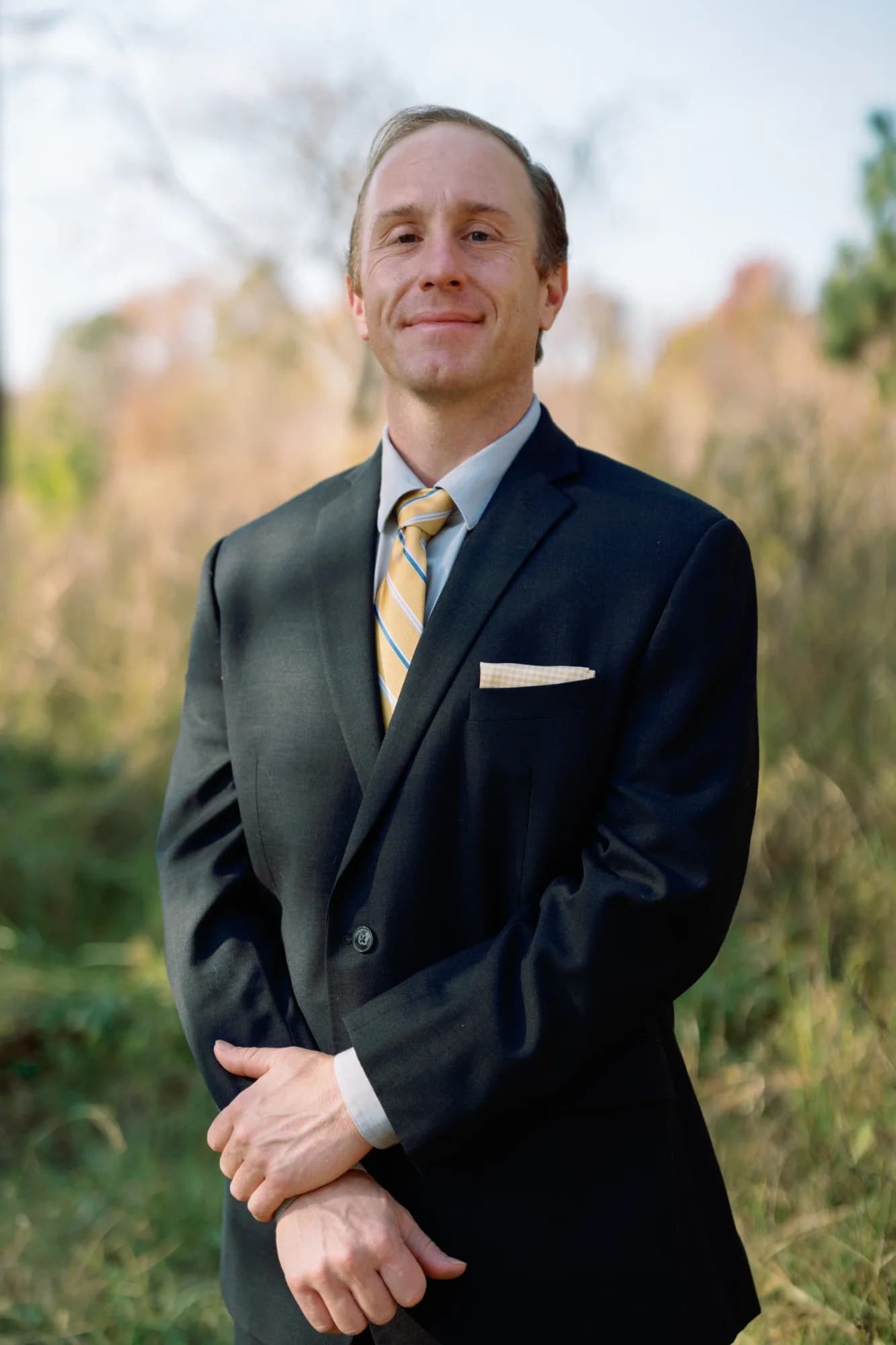 Daniel Tagge, MD, in a navy suit and gold tie, standing outdoors in tall grass