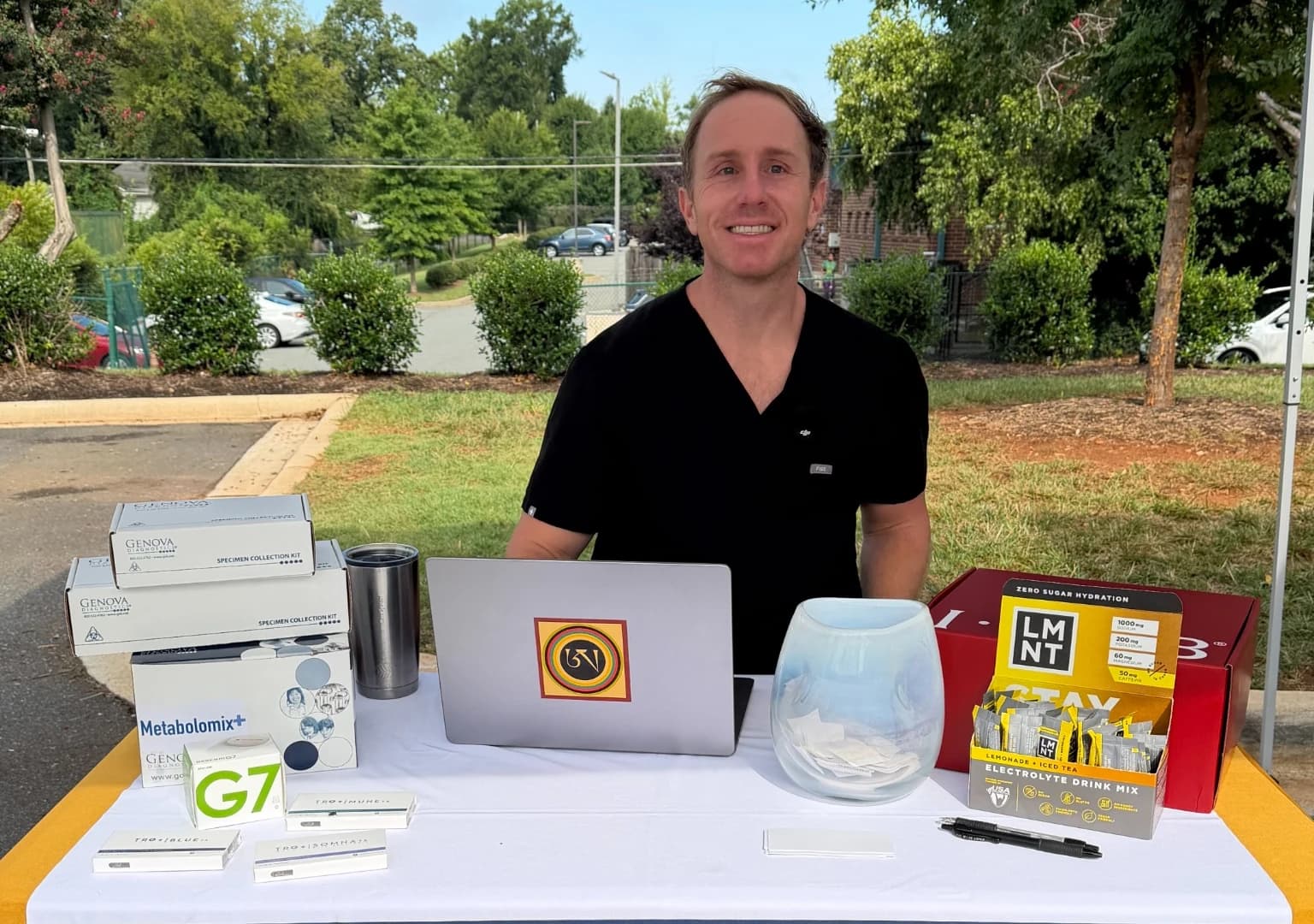 Dr. Daniel Tagge in scrubs behind a table at an outdoor community event in Charlotte, with laboratory collection kits and a laptop in front of him